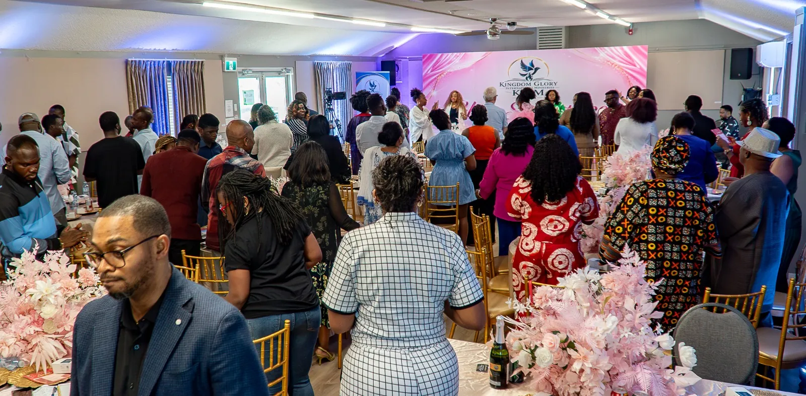 A full hall of worshippers standing together facing the stage beneath the Kingdom Glory backdrop