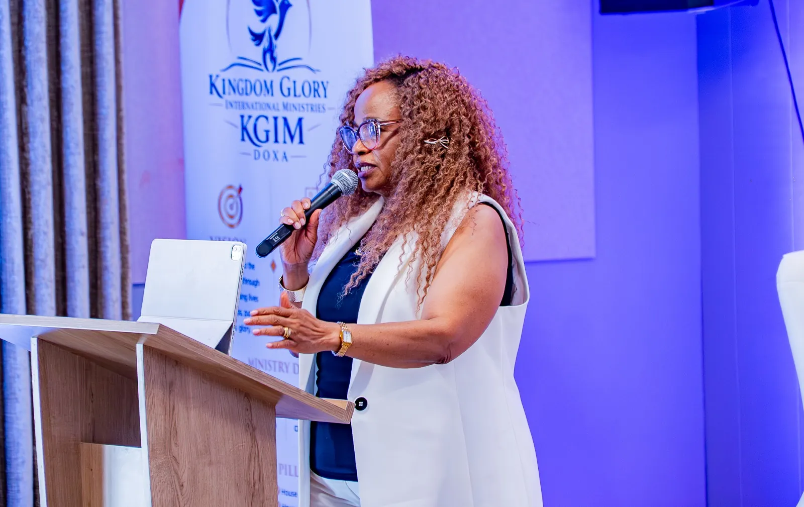 A woman speaking at a podium in front of the Kingdom Glory International Ministries banner